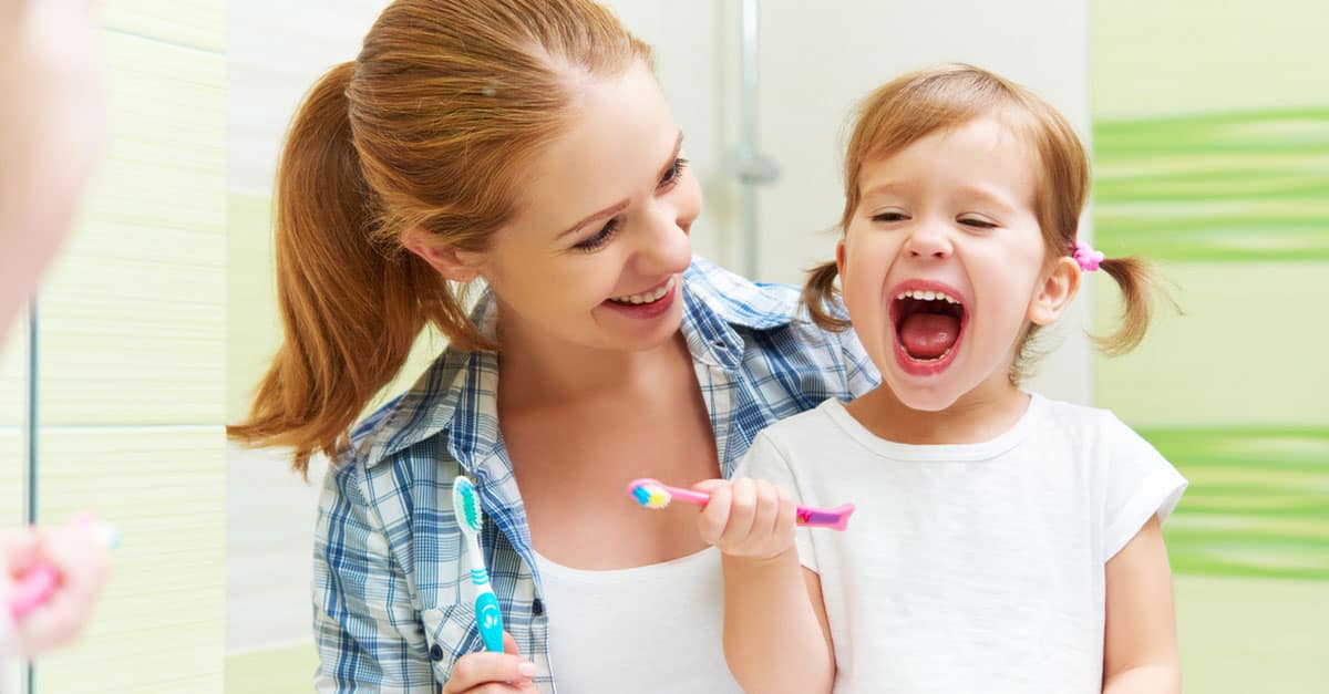A child smiles with a toothbrush in her hand as her mother looks on